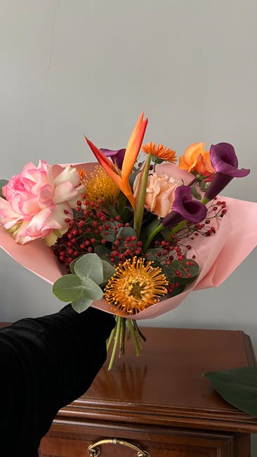 A vibrant floral bouquet featuring a mix of colorful flowers, including purple calla lilies, orange and yellow Protea, pink roses, a striking orange bird of paradise, and red hypericum berries, all ar