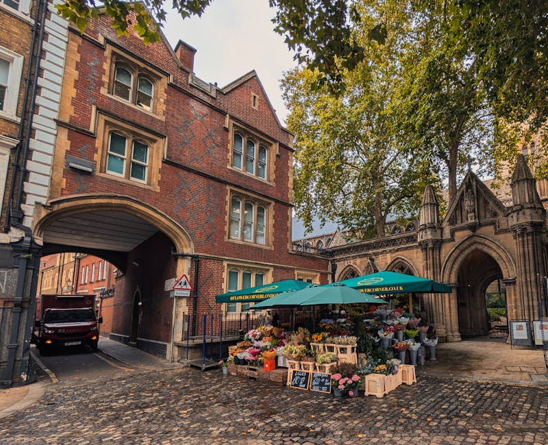 A vibrant flower market set up outdoors on a cobbled street beside a historic brick and stone building with an archway, featuring a green umbrella and wooden crates overflowing with a variety of fresh